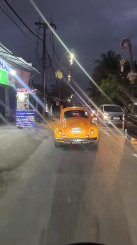 The video captures a nighttime view from a moving vehicle on a paved road. The primary focus is a bright orange Volkswagen Beetle driving ahead. The car has its brake lights illuminated. Other vehicles, including a white van and motorcycles, are visible further down the road, some with their headlights on. The environment appears to be a village or a rural area, with low-rise buildings on the left side of the road. Some of these buildings are decorated with lights, possibly for a festive occasion. Overhead, there are power lines and streetlights. The sky is dark, suggesting it is evening or night. The road surface appears wet, indicating recent rain. The overall mood is somewhat moody due to the low light conditions and the presence of streetlights and vehicle lights creating streaks of light.