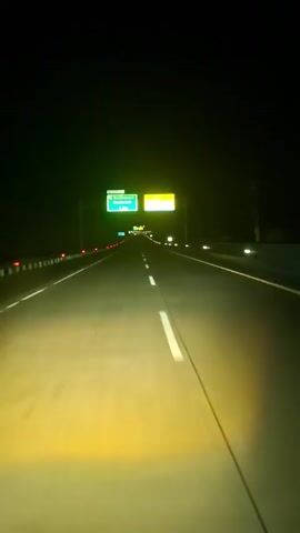 A front-view, low-angle video shows a car driving on a dark highway at night. The road is illuminated by the car's headlights, casting a yellow light on the asphalt. Two large overhead signs are visible in the distance. The left green sign indicates "Seulimeum Seulawah 1 Km" with a left arrow icon, and the right yellow sign says "Akhir Jalan Tol 1 Km" (End of Toll Road 1 Km). White dashed lines separate the lanes on the road. Red reflectors are visible along the left side of the road, and some lights are visible on the right. The sky is dark, and there are no other visible cars in the immediate vicinity. The scene is quiet, with only the sound of the car driving on the road.