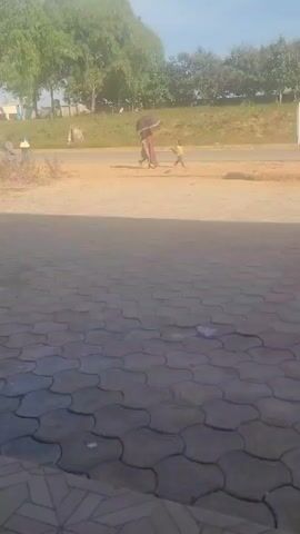 The video shows a street scene with a woman holding an umbrella and walking with a child on a dirt road. Other pedestrians walk on the side of the road, and a vehicle drives by. The foreground is a paved area with a patterned surface. In the background, there are trees and a slightly elevated green area.