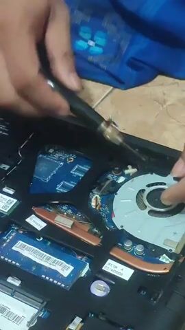 Here is a description of the video:

The video shows a person sitting on the floor, working on the inside of a laptop. The laptop is open, with its internal components visible, including the motherboard, RAM, a cooling fan, and other electronic parts. The person is using a tool, possibly a screwdriver or a specialized repair tool, to manipulate or adjust something within the laptop's interior. The camera angle is positioned above the laptop, providing a close-up view of the repair process. A blue patterned cloth is visible behind the laptop, suggesting the person might be working in a domestic setting. The lighting in the scene appears to be from overhead, illuminating the details of the laptop's components and the person's hands.
