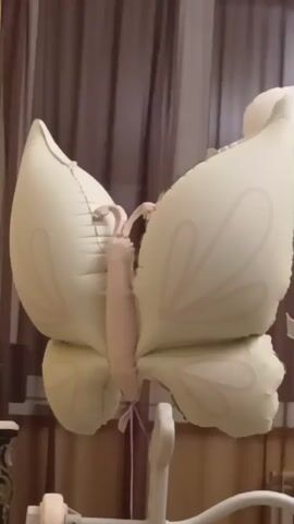A close-up shot captures a pale yellow butterfly-shaped balloon suspended by a pink ribbon against a blurred background of brown curtains and a window. The balloon is inflated and features delicate pink floral patterns on its wings. Its body is a lighter pink color, and its antennae are visible. The balloon appears to be attached to a white support structure, partially visible at the bottom of the frame. The lighting is dim, suggesting the photo was taken indoors, possibly in a bedroom.