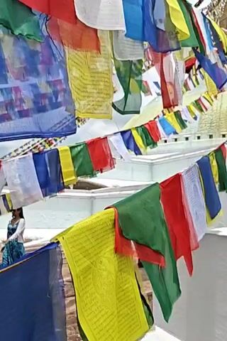 The video shows strings of colorful prayer flags fluttering in the wind. The flags are of various colors, including blue, yellow, red, green, and white, and many have Tibetan script printed on them. In the background, a white structure, possibly a stupa, is visible, with its golden spire at the very top. There are a few people walking along the white structure. One person in a pink garment walks away from the camera on the left. Further to the right, two people are visible near the edge of the white structure, one in a red shirt and another in a yellow top. The environment appears to be outdoors, with a cloudy sky overhead. The camera angle is low, looking upwards at the flags and the structure.