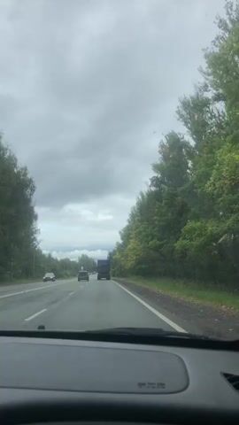 A car drives down a two-lane highway on a cloudy day. The camera is positioned from inside the car, looking forward through the windshield. Dense green trees line the right side of the road, with some showing autumn colors. A large truck and two smaller cars are visible on the road ahead, traveling in the same direction. The sky is overcast with gray clouds.
