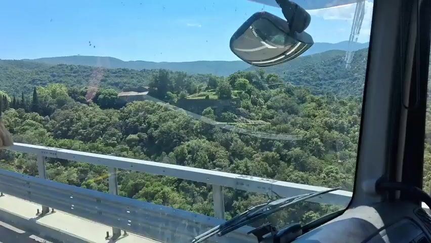 The video shows the view from the cab of a truck, looking out across a forested landscape. A building with a red-tiled roof is visible on a small hill, partially obscured by trees. The truck is traveling on a bridge, as evidenced by the safety barrier visible in the foreground. The weather is sunny and clear, with a blue sky in the background. Music is playing in the background.