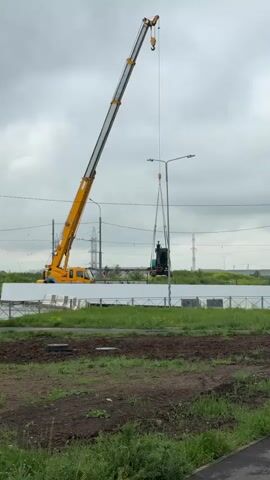A bright yellow crane is used to lift a small green excavator over a white fence. The crane is tall and the excavator is suspended by heavy chains. The surrounding environment includes a grassy area in the foreground, power lines, a road, and a cloudy sky.