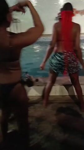 The video captures a lively poolside gathering with several individuals dancing to music. Two women in bikini tops and shorts are prominently featured dancing near the edge of a pool. The woman in the foreground wears a dark bikini top and shorts, while the other wears a black bikini top and patterned shorts with long red braids. A DJ can be seen in the background on a raised platform. Additional attendees are present, and one person is holding a tablet. The environment is festive, with clear evidence of a party or social event occurring around the pool. The lighting is dim, indicating it is evening or night.