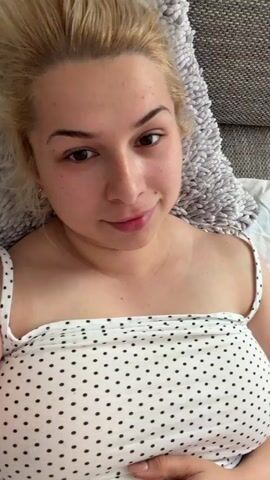 A woman with blonde hair and fair skin, wearing a white polka dot top, is lying on a couch. She is looking at the camera and speaking. The room appears to be a living room.