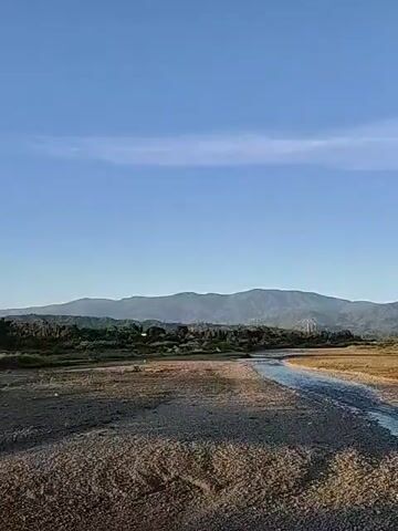 The video shows a wide river with a shallow stream of water flowing through it. The riverbed is mostly gravel and dirt, with some grassy areas on the sides. In the distance, there are mountains covered in trees. The sky is blue with a few wispy clouds. The video is static.