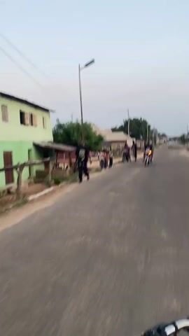 A first-person perspective from a moving vehicle shows a street scene. On the left, a street light pole stands near a green building, where several children are gathered. A man is seen walking away on the left side of the road. On the right side, a woman is seated on a black motorcycle, and a man holding crutches stands beside her, appearing to converse. Another person on a motorcycle is approaching in the distance. The road is paved and lined with buildings and sparse vegetation under an overcast sky.