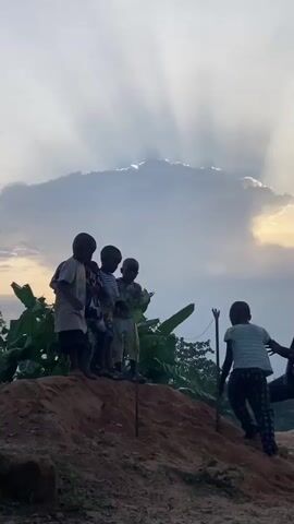 A low-angle shot shows five dark-skinned children on top of a small earthen mound at dusk. Four children stand, while one climbs up. A young child in a white shirt and black shorts jumps up and down, and a boy to his left raises both arms as well. Behind them is a cloudy sky with rays of sun streaming down. Lush, green vegetation is to the left of the mound.