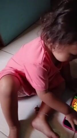 A young child wearing a pink t-shirt is sitting on the floor and looking at a smartphone. The child's left hand is touching the screen. The phone is displaying cartoon images. The floor appears to be tiled. The background is out of focus but suggests an indoor environment.