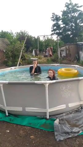 Two individuals are in an above-ground swimming pool. The person on the left, with dark hair, is wearing a black shirt and appears to be talking or gesturing. The person on the right, with reddish-brown hair, is also wearing a dark shirt and is partially submerged, looking towards the person on the left. A yellow inflatable ring is floating in the pool near the person on the right. The pool is surrounded by a dirt area with some debris. In the background, there are trees, a building with a corrugated roof, and a clothesline with laundry hanging. The sky is overcast. The individuals seem to be interacting playfully.