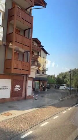 The video shows a view from a vehicle, likely a bus, moving along a road. On the left is a multi-story building with wooden balconies and brick accents, possibly a hotel or apartment building. A sign on the building reads "CENTRO COMMERCIALE" and "FARMACIA". In front of the building is a small parking area with several vehicles, including black cars, a white van, and a silver mini-van. To the right of the parking area is a grassy hill with trees. The road curves to the right, and the view transitions to a wider shot of the grassy hills and trees. The sky is clear and blue. The lighting suggests it is daytime, possibly morning or late afternoon due to the long shadows.