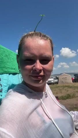 A woman is sitting in a chair outdoors on a sunny day. She has a small plant growing out of the top of her head. In the background, there are tents, cars, and a body of water. The woman is looking at the camera and smiling.