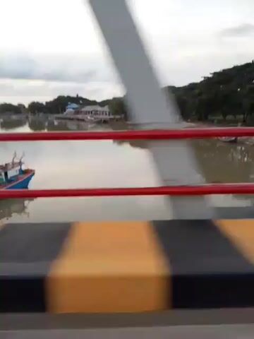 The video shows a first-person perspective traveling across a bridge. The bridge has a metal truss structure overhead and red railings along the sides. The road surface is visible, with yellow and black striped barriers along the edge. Below the bridge, a body of water is visible, with several small boats moored along the shore. Trees line the far bank of the water, and in the distance, there appear to be some buildings. The sky is overcast. The footage appears to be taken from a vehicle, as evidenced by the motion blur and the perspective.