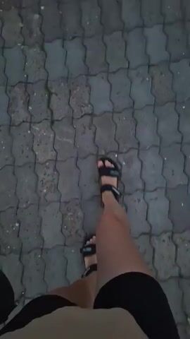 A person is walking on a paved walkway. They are wearing black shorts and black sandals. The walkway is made of interlocking gray pavers. The camera is pointed down at the person's legs and feet as they walk. The lighting is dim, suggesting it is either early morning or late evening.