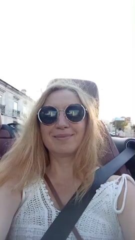 A woman with blonde hair and sunglasses smiles as she sits in the driver's seat of a convertible. The sun is bright, and there are buildings and trees in the background. She is wearing a white top and a dark seatbelt.