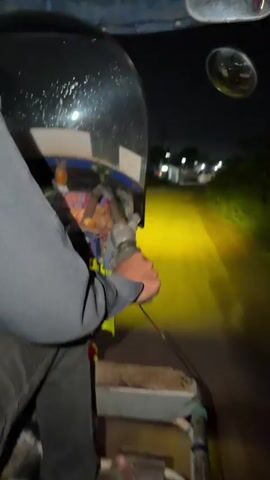 This media file captures a first-person perspective from what appears to be the back of a three-wheeled vehicle, likely a tuk-tuk, traveling at night on a road in Thakhek, Laos. The primary subject is the hand of the person operating the vehicle, gripping a handle. The vehicle itself is partially visible, including a portion of its dark-colored roof and some decorative items.

The setting is a rural or semi-rural road illuminated by the vehicle's bright yellow headlights, casting a strong glow on the asphalt ahead. The surrounding environment is dark, with the faint outlines of trees and vegetation visible on the sides of the road. In the distance, a few scattered lights suggest the presence of houses or small settlements. The time of day is clearly night, indicated by the darkness and the artificial illumination.

There is no discernible weather beyond the darkness. The mood conveyed is one of movement and perhaps a sense of quiet travel through the night. The focus is on the act of driving and the immediate surroundings.

Visible text is minimal. On the roof of the vehicle, a white sticker with some unreadable text is present. The overall impression is a candid glimpse into nighttime travel in a Southeast Asian setting.