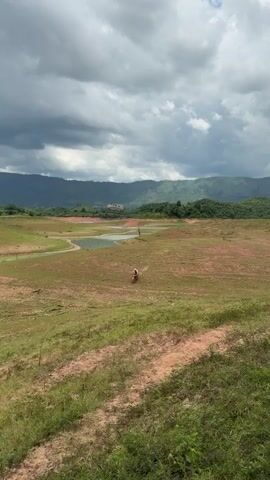 A person rides a dirt bike across a wide, dry, grassy landscape. The rider is visible in the middle distance, wearing a helmet and a light-colored shirt. The dirt bike is red. The terrain is mostly dry and brown, with patches of green grass. In the background, there is a body of water and rolling hills covered in trees. The sky is cloudy and dramatic, with dark clouds dominating the upper portion of the frame. A dirt path is visible in the foreground.