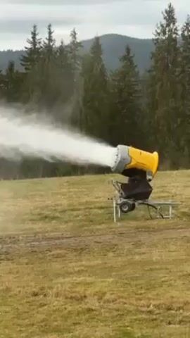 The video shows a yellow snow cannon in a grassy field. The cannon is pointed towards the left side of the frame and is actively spraying a mist of water into the air. Behind the cannon are several tall evergreen trees and a mountain range can be seen in the distance. The sky is overcast. The ground around the cannon is a mix of grass and muddy patches. The snow cannon is labeled "TT10". There is background sound of someone speaking.