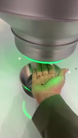 A person wearing a green jacket holds their hand under a metal container, which emits a green light. When the light turns off, the person shows a chunk of dark chocolate. The scene is lit by overhead lights, and the container sits on a white table. A small metal bowl is on the table under the metal container.