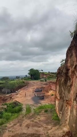 The video shows a quarry landscape under a cloudy sky. The scene features a large, brown rock cliff on the right and a person standing in the middle of a dirt road. A dump truck drives up the road, emitting a cloud of black smoke. In the background, there is a green landscape with trees and a view of a distant town.