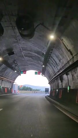 The video shows a point-of-view perspective of driving through a tunnel and emerging onto a highway. The tunnel has a concrete arched ceiling with lights and traffic signals visible. The traffic signals display green arrows and a speed limit of 80. As the vehicle exits the tunnel, the scene opens up to a highway with multiple lanes. The time of day appears to be either dawn or dusk, with a soft, warm light illuminating the sky. The sky is partly cloudy, and in the distance, hills or mountains are visible. The weather seems clear and calm. The activity is driving, transitioning from an enclosed, artificial environment to an open, natural landscape. There are no people visible in the video. Objects include the road, tunnel structure, traffic lights, signs, street lamps, guardrails, and a truck in the distance. The location is implied to be Praveshka Lakavitsa, Bulgaria.