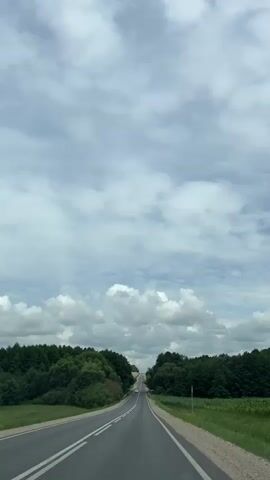 A vertical shot captures a straight, paved road stretching towards the horizon under a cloudy sky. Lush green trees line both sides of the road, with a hint of farmland visible on the right. A white car is seen driving on the road, passing by trees. The scene appears to be an outdoor, daytime shot.