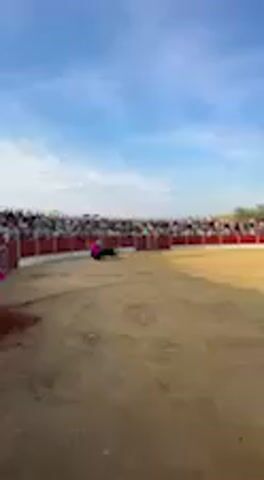 This video captures a bullfighting event within an outdoor bullring in San José del Valle, Spain. A bullfighter, wearing traditional bullfighting attire, is seen in the sandy arena engaging with a dark-colored bull. The bullfighter skillfully uses a large, brightly colored cape, which is pink on one side and yellow/orange on the other, to direct the bull's movements, performing a series of passes. The bull responds to the cape, charging and turning.

The bullring is enclosed by a red barrier, and behind it, a large audience fills the stands, observing the spectacle. The sky overhead is bright with scattered clouds, indicating that the event is taking place during the day. On a section of the red barrier to the right of the bullfighter, a prominent white number "6" is visible. The atmosphere suggests an engaged crowd at this traditional cultural event.