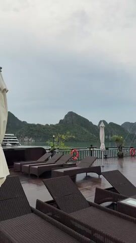 The video displays a serene scene from the deck of a boat in Hải Phòng, Vietnam. The foreground is dominated by rows of dark brown wicker lounge chairs and small glass-topped tables, suggesting a relaxed, resort-like atmosphere on the vessel. The wooden deck appears damp, hinting at recent rain or sea spray.

In the mid-ground, several people are visible. Some are seated or reclining on the lounge chairs, possibly enjoying the view or resting. Others are standing and interacting near a white structure that resembles a bar or lounge area on the boat, with some individuals appearing to be staff. A white umbrella is also present, partially obscuring the bar area. Two orange life preservers are mounted on the railing, a common safety feature on boats.

The background showcases the breathtaking scenery of Halong Bay. Towering limestone karsts covered in lush greenery rise dramatically from the calm, green water. The sky is overcast, suggesting a cloudy or potentially rainy day, but the diffused light contributes to a peaceful ambiance. The overall mood is tranquil and scenic, highlighting the natural beauty of the location. The image captures a moment of leisure and natural wonder.