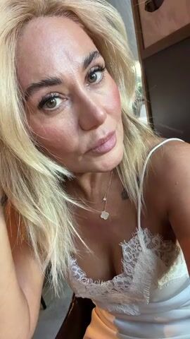 The video shows a close-up, head-and-shoulders shot of a woman with blonde hair and fair skin. She is wearing a white, lacy camisole and a delicate necklace with a clover-shaped pendant. Her left hand is resting on her chin, and she has a tattoo on her wrist that appears to say "Belle". The woman's makeup is visible, including blush on her cheeks and mascara on her eyelashes. Her eyes are looking downwards, and her lips are slightly pursed. The background is out of focus, but appears to be an indoor setting with warm lighting. The video is approximately four seconds long and consists of several still frames that appear to be taken in rapid succession, creating a slight flickering effect.