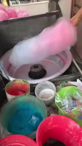 The video shows a person making cotton candy.  The person's hand holds a large, pink-and-white cloud of cotton candy above a metal machine with a rotating center.  Around the machine are several containers filled with colored sugar, including red, blue, and white. Some pre-made pink cotton candy in bags is visible in the background.