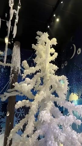 A video shows a white, frosted Christmas tree in an indoor setting. Icicles hang from its branches, and snow is visible at its base. To the left, a brown wooden pole is also covered in frost and icicles. The background features a dark blue wall with a starry sky design, snowflakes, and stylized white trees. Lights are visible overhead, and a railing is present in the foreground. The video is static, with no people or movement observed.