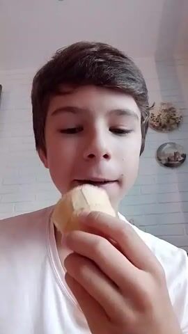 A close-up shot shows a young person in a white t-shirt. The person is holding a vanilla ice cream cone in their right hand and eating it. Their mouth is open as they take a bite of the ice cream. The background appears to be a wall with some decorative plates.