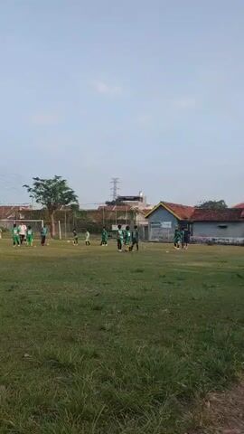 A soccer game is being played on a grass field. The sky is clear and blue, with a few clouds. There are several people on the field, all wearing green uniforms. The people are spread out across the field, and some of them are kicking a soccer ball. There are also some buildings and trees in the background.