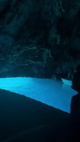 The video shows the interior of a sea cave with strikingly blue water. In the foreground, the silhouetted head and shoulders of a person facing to the right are visible. They appear to be looking at a phone held in their hand. To the left, a large expanse of intensely blue water reflects the cave's illumination. In the middle ground, a white boat with passengers is visible, and lettering on the side of the boat reads "CENTAR KOMIŽA". The cave walls are dark and textured, with various rock formations visible. Another person is seen standing in the boat, looking towards the cave's interior. The lighting in the cave creates a surreal, glowing effect on the water. The scene suggests a tourist excursion into a natural cave.