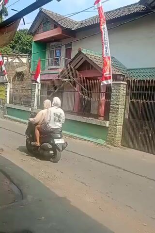 A motorcycle carrying two people rides down a street. The person in front is wearing a dark jacket and a helmet, while the person in the back is wearing a light-colored shirt and a headscarf. They are riding past a building with an Indonesian flag displayed on it. The building is painted green and pink with a white fence. Trees are visible in the background. The street is paved and has a curb on the left side. A yellow banner with text in Indonesian is hanging on the left side of the frame.