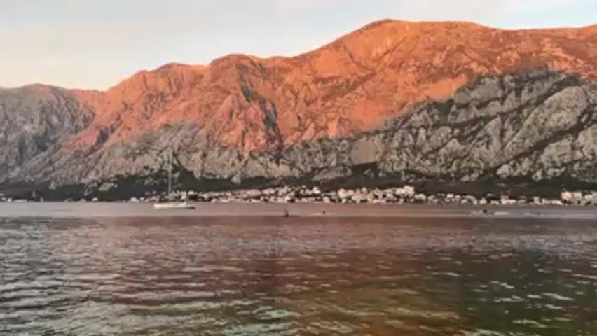 The video shows a body of water with mountains in the background.  The mountains are mostly brown and rocky, but have a strip of green at the base. The sun is low, casting a warm glow on the mountains. A small town is situated along the base of the mountains at the water’s edge. There is a sailboat in the water, as well as what appear to be several people swimming.