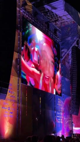The video begins with a close-up of a large LED screen displaying a distorted image of a person's face, with vibrant lights and colors. The screen is mounted on a metal scaffolding structure, with speakers on either side. Below the screen, a crowd of people can be seen, illuminated by the stage lights. The camera then pans to the left, showing more of the scaffolding and a banner with logos. The video then briefly cuts to a blurry shot of the stage and the crowd from a different angle, with more lights and movement. Finally, the scene transitions to a shot of a crowd of people in darkness, with a street lamp visible in the background. Some people are holding up their phones, likely recording the event.