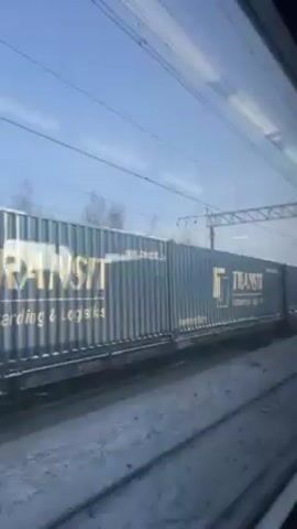 The video shows a view from inside a train, looking out the window at another train moving in the opposite direction. The other train is a freight train carrying shipping containers. The containers are branded with "TRANSIT Forwarding & Logistics" and are colored blue and red. The ground is covered in snow. The sky is clear and blue. Overhead power lines are visible.