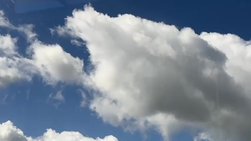 The video shows a dynamic sky filled with various types of clouds against a clear blue background. The clouds are predominantly white and puffy, with some darker, more textured formations appearing to be developing or in the process of dissipating. There's a sense of movement within the clouds, suggesting wind. In the upper left corner of the frame, a sliver of what appears to be a structure, possibly a window frame or part of a building, is visible, indicating the perspective is from inside or through a structure looking out. The lighting suggests it is daytime, with sunlight illuminating the cloud formations.