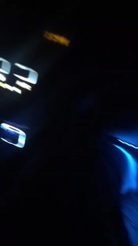 The video shows a car driving on a dark, winding road at night. The headlights illuminate the road ahead, and the dashboard lights are visible in the foreground. The road is paved with a white line marking the center. There is green vegetation lining the sides of the road. Another vehicle can be seen approaching in the distance. The video is somewhat blurry.