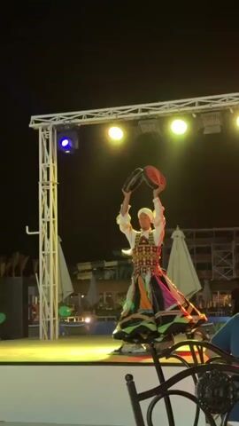 Here is a description of the video:

The video shows a man performing a traditional dance on a brightly lit stage at night. He is wearing a colorful, layered skirt, a white shirt, an embroidered vest, and a white turban. He is holding two round, dark objects in his hands, possibly tambourines, and moves them in time with the music. The stage is illuminated with spotlights of varying colors, including purple, green, and white. Behind the stage are various structures and objects, including white umbrellas and water park features. The sky is dark, and some foreground objects, such as chairs and people, are partially visible, suggesting the performance is being viewed by an audience.