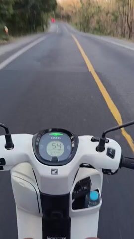 The video is a first-person perspective of someone riding a white Honda scooter on a paved road. The road has a yellow line down the center and a white line on the right edge. The rider's hands are visible on the handlebars. The scooter's digital display shows the speed decreasing from 53 to 47. The environment is outdoors with trees and hills visible on either side of the road. The lighting suggests it is daytime, possibly late afternoon due to the warm light on the trees. A red car is visible in the distance on the road ahead. A water bottle is clipped to the scooter.