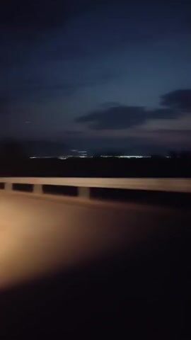 The video shows a view from a car at dusk or night. The sky is dark blue with some clouds. In the distance, there are lights from what appears to be a town or city. A guardrail runs along the side of the road in the foreground. The road itself is dark, with the headlights of the car illuminating a patch of the road. The perspective suggests the car is moving.