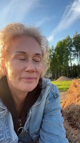 The video opens with a close-up of a woman with blonde, curly hair, pulled into a bun. She wears a denim jacket over a black top and a small hoop earring, and is looking slightly upwards and to her right with a gentle smile. The warm, golden light suggests late afternoon or early evening.

The camera then pans to show a rural outdoor setting with large piles of hay and a line of green trees in the background under a blue sky with some wispy clouds. Another woman, with long reddish-brown hair, wearing a pink bomber jacket, light-wash denim shorts, and pink and white sneakers, is seen from behind. She is actively climbing onto one of the large hay bales, using her hands and feet to navigate the loose hay. She appears focused on her ascent. The ground is grassy and uneven in the background.