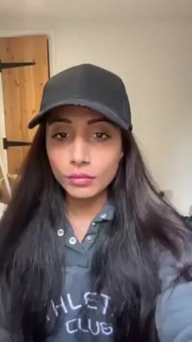 A woman wearing a black baseball cap and a grey hoodie is applying blush to her cheeks with a pink-handled brush. She looks directly at the camera periodically. She has long dark hair. In the background, a wooden door with black hardware is visible, along with part of a bed and a cardboard box.