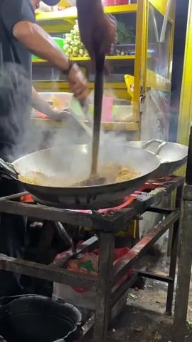 A person wearing a dark shirt and a cap is stir-frying food in a large wok over an open flame. The food appears to be noodles or rice and is emitting a significant amount of steam. The person is using two utensils, a spatula and a pair of tongs, to toss and stir the food. Another person's arm is visible in the background, also holding a utensil, suggesting they might be assisting or cooking in another wok. The scene appears to be a street food stall, with shelves stocked with ingredients and colorful containers in the background. The lighting is warm and appears to be artificial, suggesting it's nighttime or indoors. The overall atmosphere is one of busy food preparation.