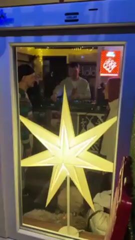 A man wearing a gray jacket over a white t-shirt is working a DJ mixer, as seen through a window. In the foreground is a lit up white star decoration. Another man wearing a striped shirt is standing next to the window, looking at the DJ. A sign for “kola” is on the wall behind the DJ. There are also posters on the wall behind the DJ.