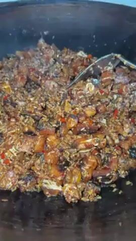 The video shows a large, dark wok containing a mixture of fried rice, diced pork, and red chilies. The food is being stirred with a large metal spatula. Steam rises from the hot dish.